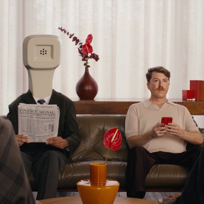 A man with a telephone handset for a head reads the paper on a sofa beside a man holding a phone