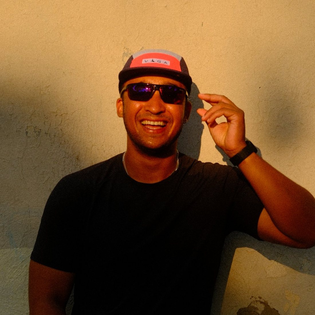 Luke AG in a cap and sunglasses against a sunlit wall, laughing